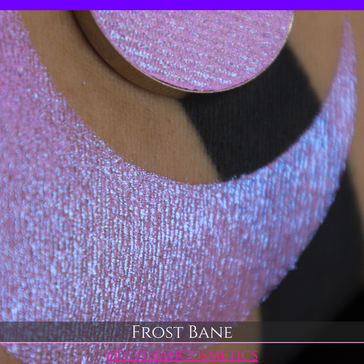 A close-up of skin with a large iridescent pink-blue crescent-shaped makeup swatch and a matching circulareyeshadow pan, with text reading 'FROST BANE' and '#DEVINAH COSMETICS' at the bottom.
