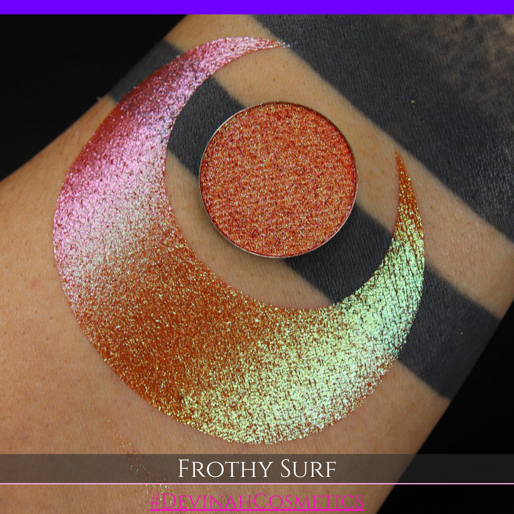 A round eyeshadow pan labeled 'FROTHY SURF' made by Devinah Cosmetics, placed over a large crescent moon swatch of the same shimmering shifty eyeshadow on skin. The eyeshadow has a duochrome multichrome shift between colors green, yellow, gold, orange, pink, red.