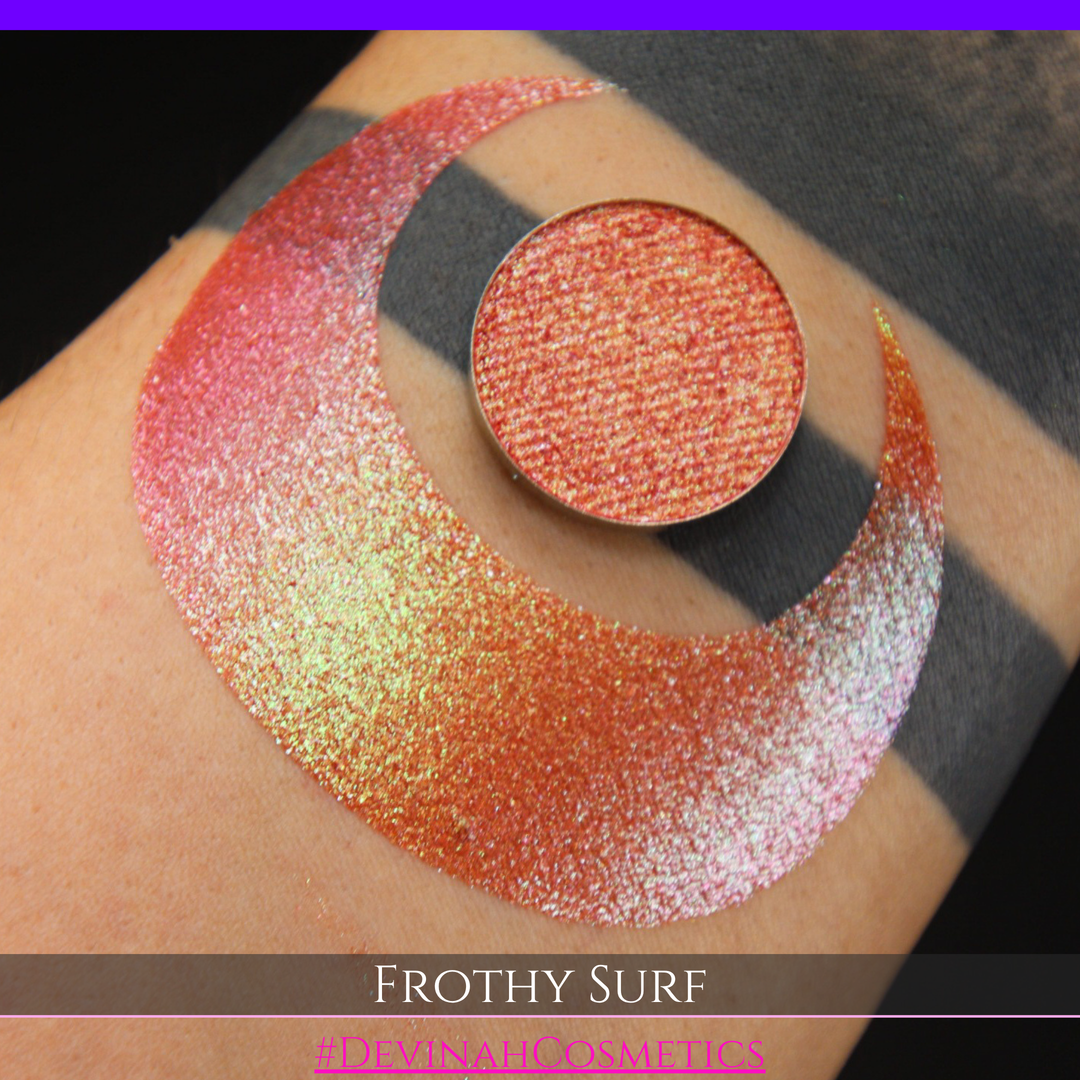 A round eyeshadow pan labeled 'FROTHY SURF' made by Devinah Cosmetics, placed over a large crescent moon swatch of the same shimmering shifty eyeshadow on skin. The eyeshadow has a duochrome multichrome shift between colors green, yellow, gold, orange, pink, red.