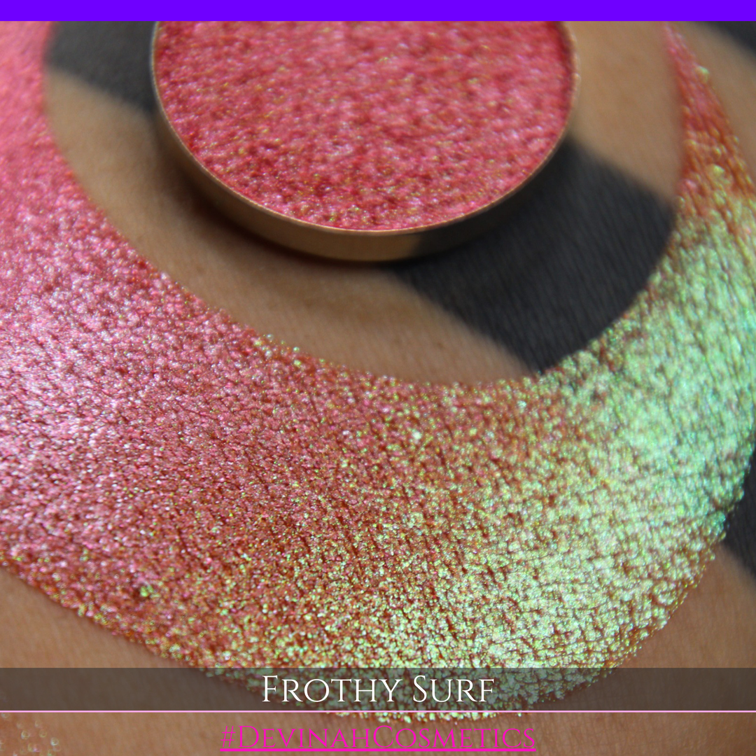 A round eyeshadow pan labeled 'FROTHY SURF' made by Devinah Cosmetics, placed over a large crescent moon swatch of the same shimmering shifty eyeshadow on skin. The eyeshadow has a duochrome multichrome shift between colors green, yellow, gold, orange, pink, red.