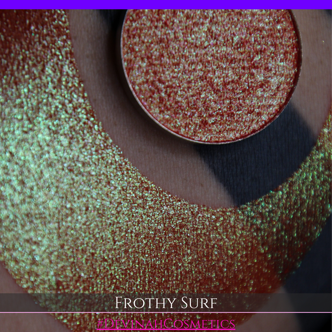 A round eyeshadow pan labeled 'FROTHY SURF' made by Devinah Cosmetics, placed over a large crescent moon swatch of the same shimmering shifty eyeshadow on skin. The eyeshadow has a duochrome multichrome shift between colors green, yellow, gold, orange, pink, red.