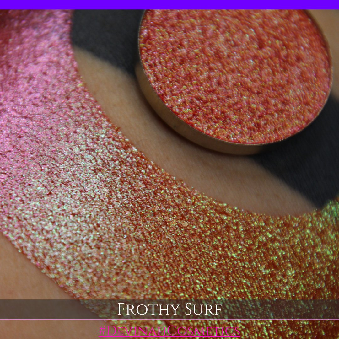 A round eyeshadow pan labeled 'FROTHY SURF' made by Devinah Cosmetics, placed over a large crescent moon swatch of the same shimmering shifty eyeshadow on skin. The eyeshadow has a duochrome multichrome shift between colors green, yellow, gold, orange, pink, red.