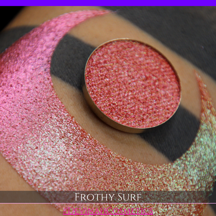 A round eyeshadow pan labeled 'FROTHY SURF' made by Devinah Cosmetics, placed over a large crescent moon swatch of the same shimmering shifty eyeshadow on skin. The eyeshadow has a duochrome multichrome shift between colors green, yellow, gold, orange, pink, red.