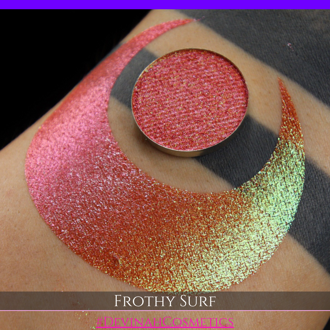 A round eyeshadow pan labeled 'FROTHY SURF' made by Devinah Cosmetics, placed over a large crescent moon swatch of the same shimmering shifty eyeshadow on skin. The eyeshadow has a duochrome multichrome shift between colors green, yellow, gold, orange, pink, red.