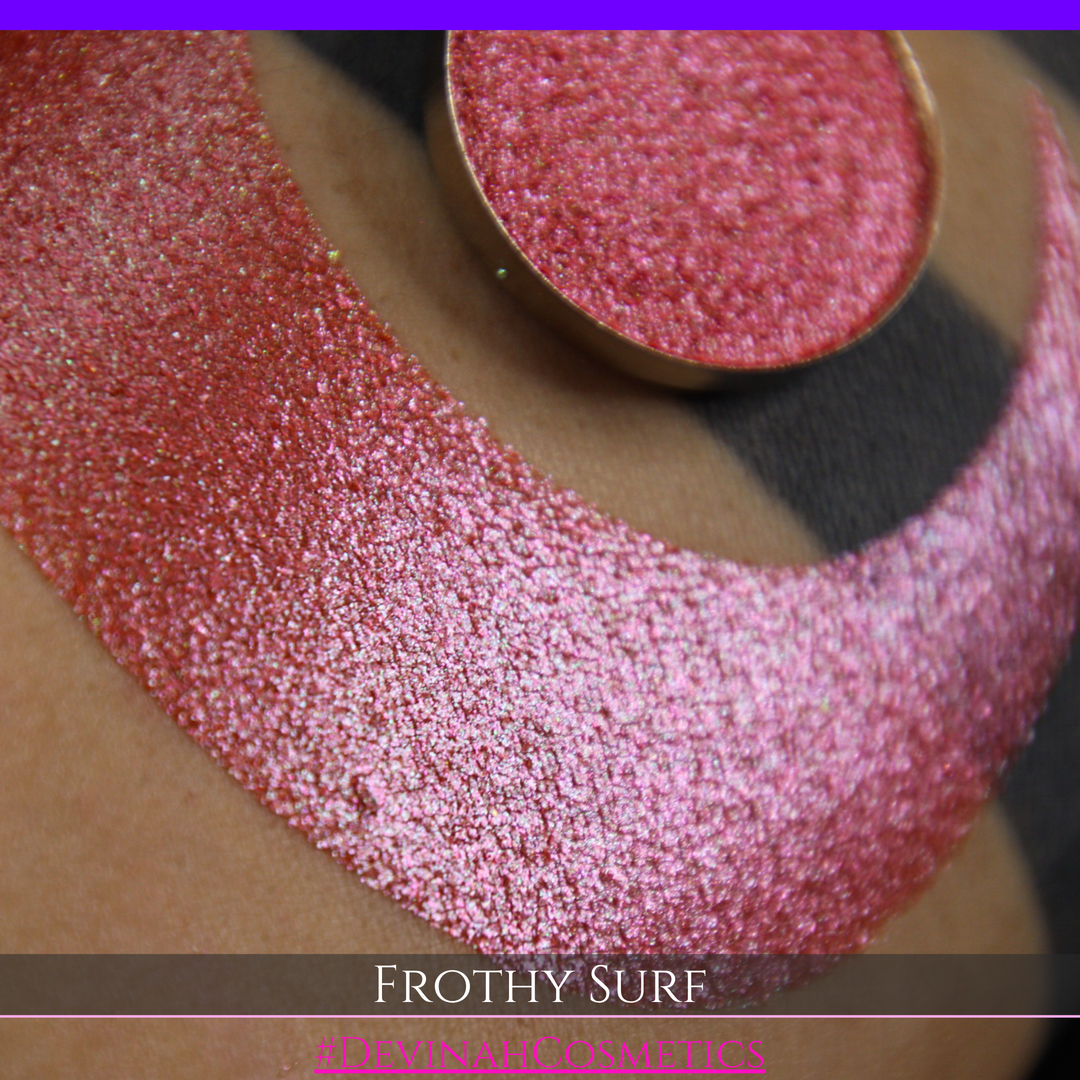 A round eyeshadow pan labeled 'FROTHY SURF' made by Devinah Cosmetics, placed over a large crescent moon swatch of the same shimmering shifty eyeshadow on skin. The eyeshadow has a duochrome multichrome shift between colors green, yellow, gold, orange, pink, red.
