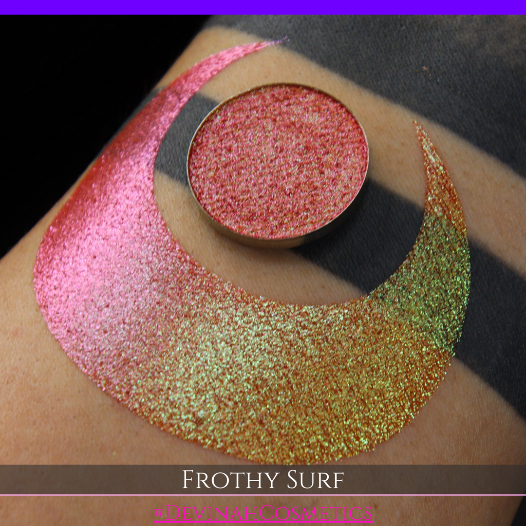 A round eyeshadow pan labeled 'FROTHY SURF' made by Devinah Cosmetics, placed over a large crescent moon swatch of the same shimmering shifty eyeshadow on skin. The eyeshadow has a duochrome multichrome shift between colors green, yellow, gold, orange, pink, red.