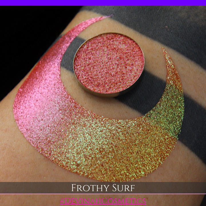 A round eyeshadow pan labeled 'FROTHY SURF' made by Devinah Cosmetics, placed over a large crescent moon swatch of the same shimmering shifty eyeshadow on skin. The eyeshadow has a duochrome multichrome shift between colors green, yellow, gold, orange, pink, red.