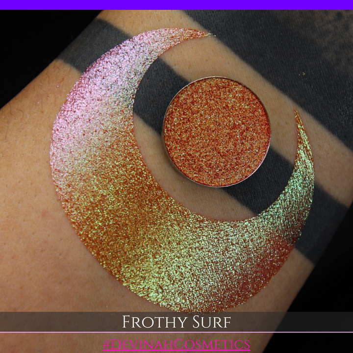 A round eyeshadow pan labeled 'FROTHY SURF' made by Devinah Cosmetics, placed over a large crescent moon swatch of the same shimmering shifty eyeshadow on skin. The eyeshadow has a duochrome multichrome shift between colors green, yellow, gold, orange, pink, red.