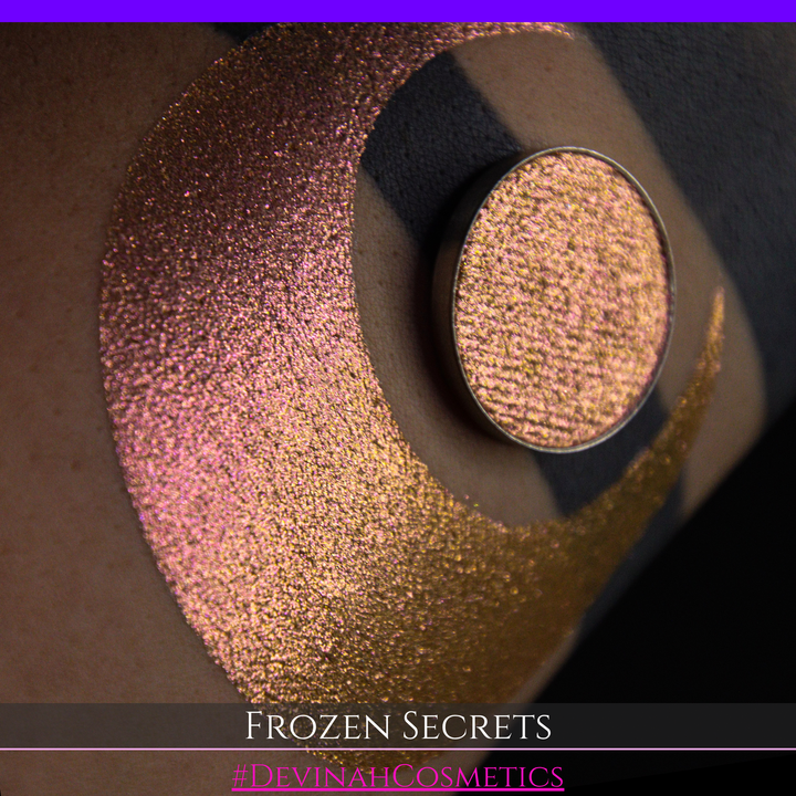A close-up of a person's arm with a large red pink-to-gold crescent moon eyeshadow makeup swatch, alongside a round eyeshadow pan. Text at the bottom reads 'FROZEN SECRETS' and '#DEVINAH COSMETICS'.
