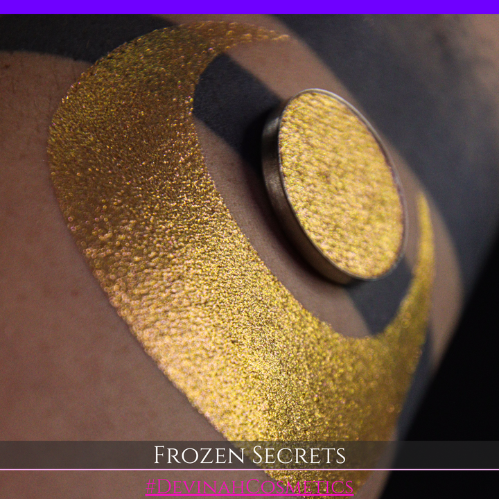 A close-up of a person's arm with a large red pink-to-gold crescent moon eyeshadow makeup swatch, alongside a round eyeshadow pan. Text at the bottom reads 'FROZEN SECRETS' and '#DEVINAH COSMETICS'.