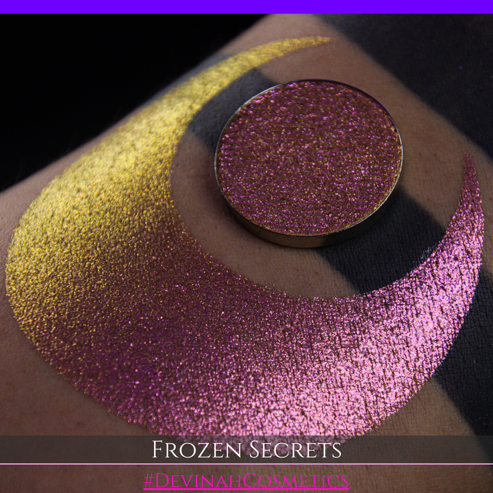 A close-up of a person's arm with a large red pink-to-gold crescent moon eyeshadow makeup swatch, alongside a round eyeshadow pan. Text at the bottom reads 'FROZEN SECRETS' and '#DEVINAH COSMETICS'.