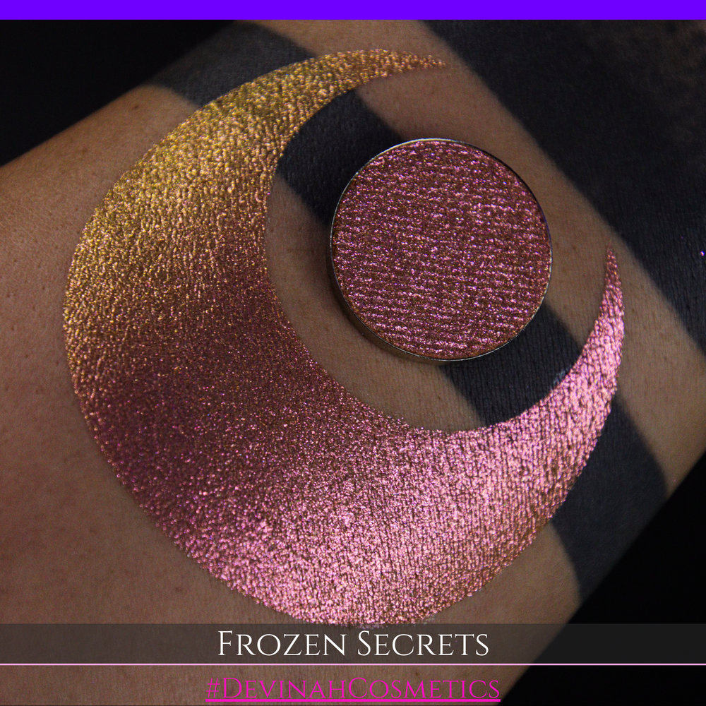 A close-up of a person's arm with a large red pink-to-gold crescent moon eyeshadow makeup swatch, alongside a round eyeshadow pan. Text at the bottom reads 'FROZEN SECRETS' and '#DEVINAH COSMETICS'.
