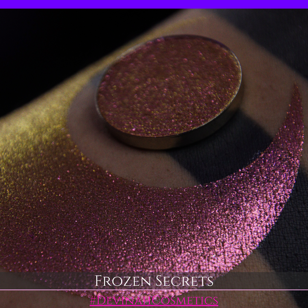 A close-up of a person's arm with a large red pink-to-gold crescent moon eyeshadow makeup swatch, alongside a round eyeshadow pan. Text at the bottom reads 'FROZEN SECRETS' and '#DEVINAH COSMETICS'.