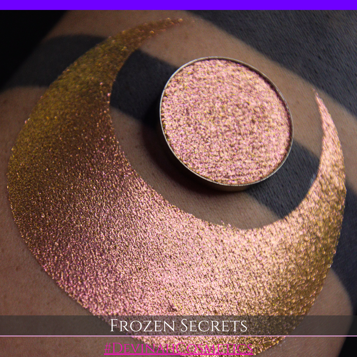 A close-up of a person's arm with a large red pink-to-gold crescent moon eyeshadow makeup swatch, alongside a round eyeshadow pan. Text at the bottom reads 'FROZEN SECRETS' and '#DEVINAH COSMETICS'.