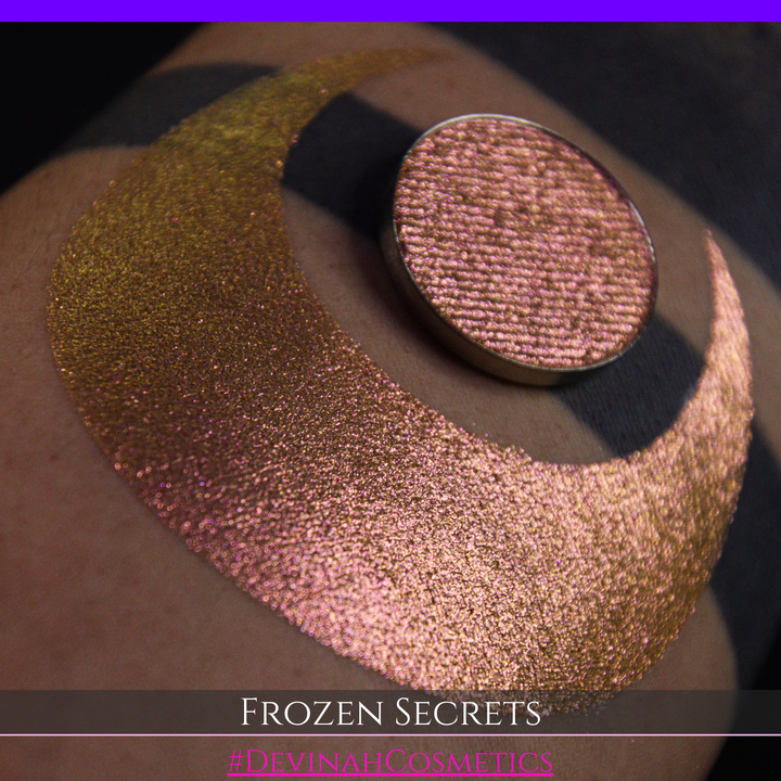 A close-up of a person's arm with a large red pink-to-gold crescent moon eyeshadow makeup swatch, alongside a round eyeshadow pan. Text at the bottom reads 'FROZEN SECRETS' and '#DEVINAH COSMETICS'.