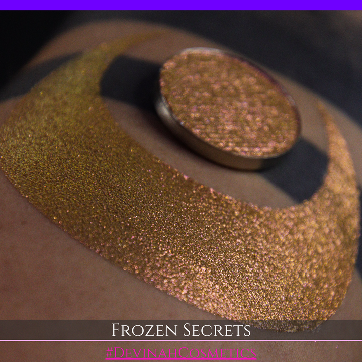 A close-up of a person's arm with a large red pink-to-gold crescent moon eyeshadow makeup swatch, alongside a round eyeshadow pan. Text at the bottom reads 'FROZEN SECRETS' and '#DEVINAH COSMETICS'.