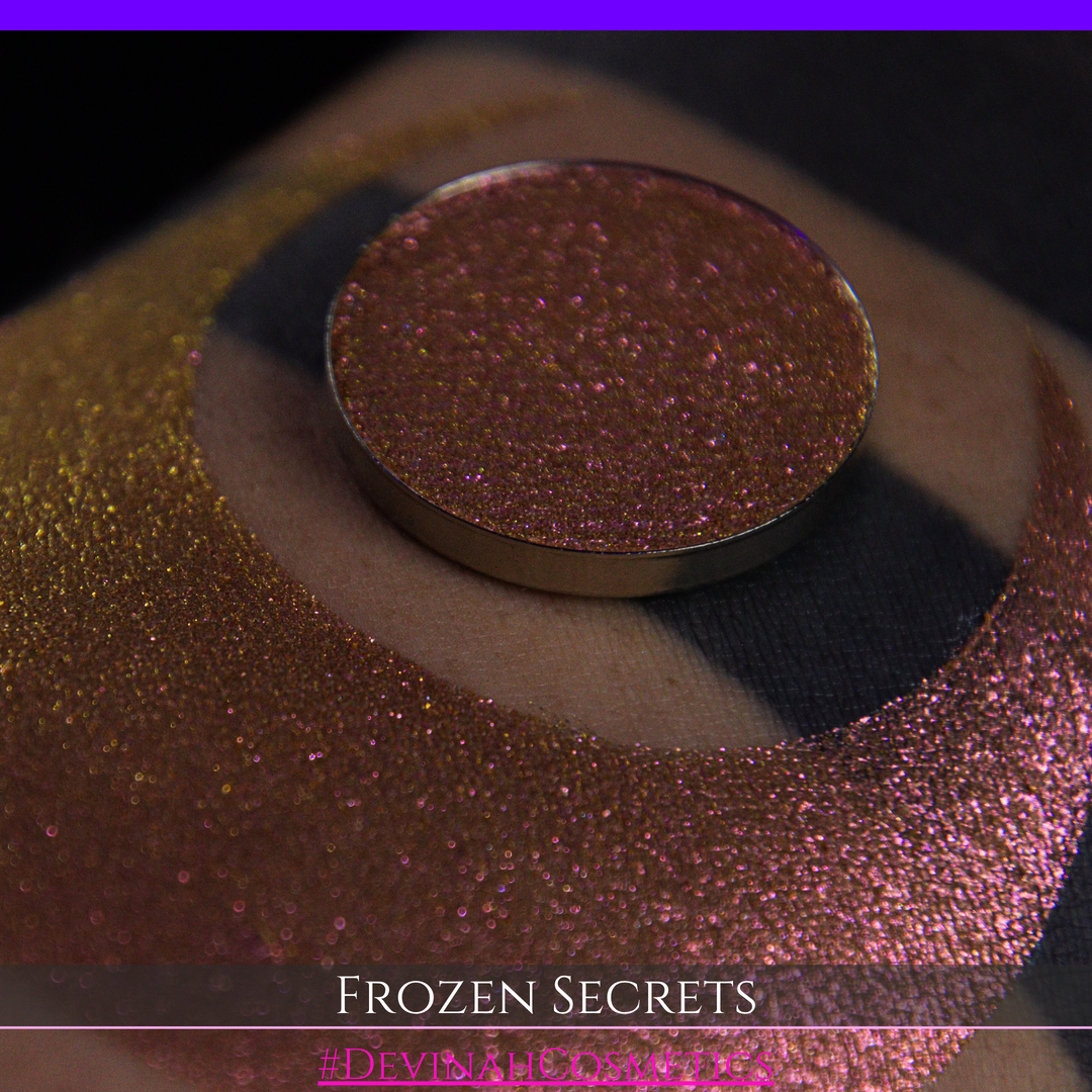 A close-up of a person's arm with a large red pink-to-gold crescent moon eyeshadow makeup swatch, alongside a round eyeshadow pan. Text at the bottom reads 'FROZEN SECRETS' and '#DEVINAH COSMETICS'.