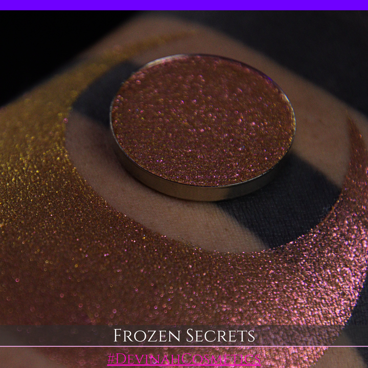 A close-up of a person's arm with a large red pink-to-gold crescent moon eyeshadow makeup swatch, alongside a round eyeshadow pan. Text at the bottom reads 'FROZEN SECRETS' and '#DEVINAH COSMETICS'.