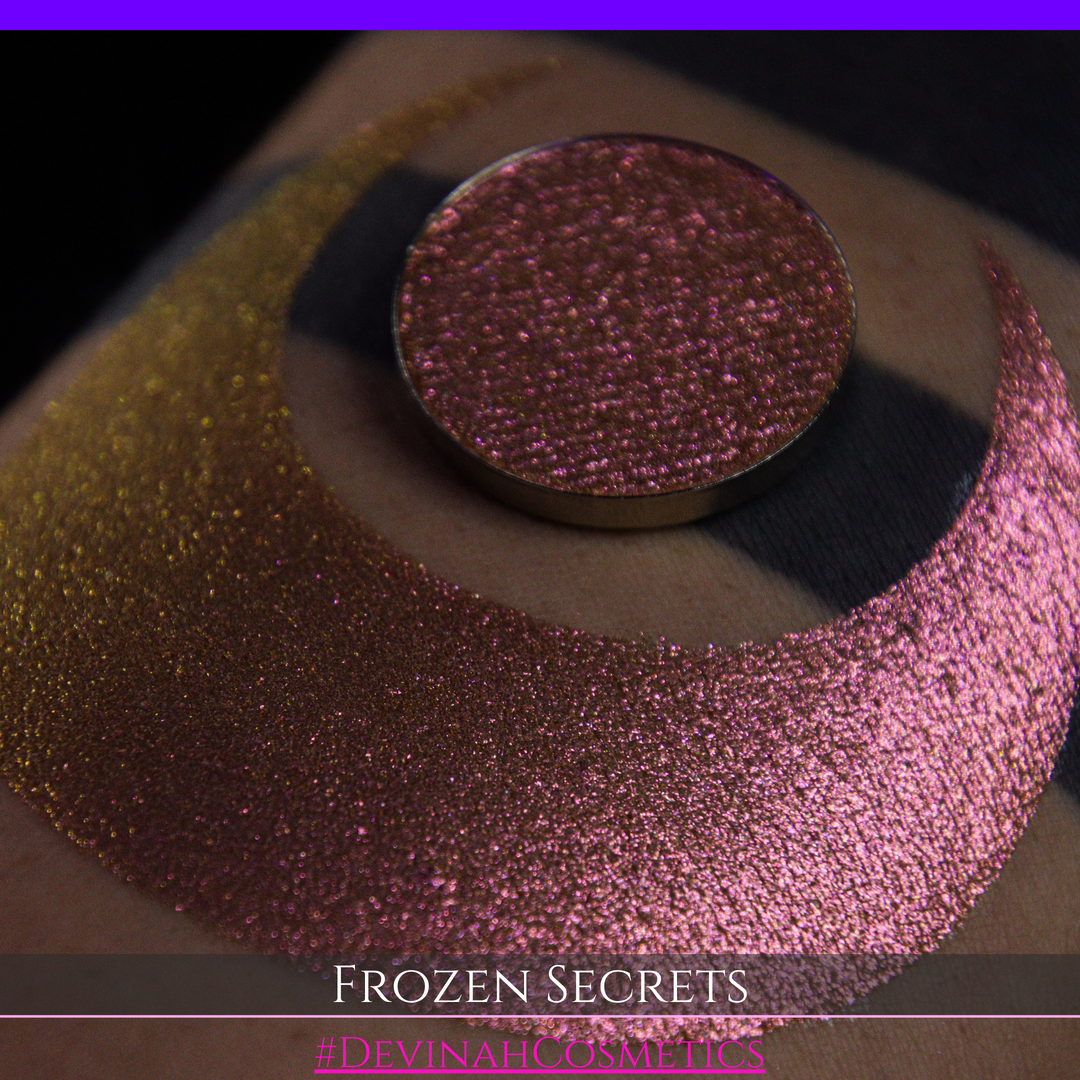 A close-up of a person's arm with a large red pink-to-gold crescent moon eyeshadow makeup swatch, alongside a round eyeshadow pan. Text at the bottom reads 'FROZEN SECRETS' and '#DEVINAH COSMETICS'.