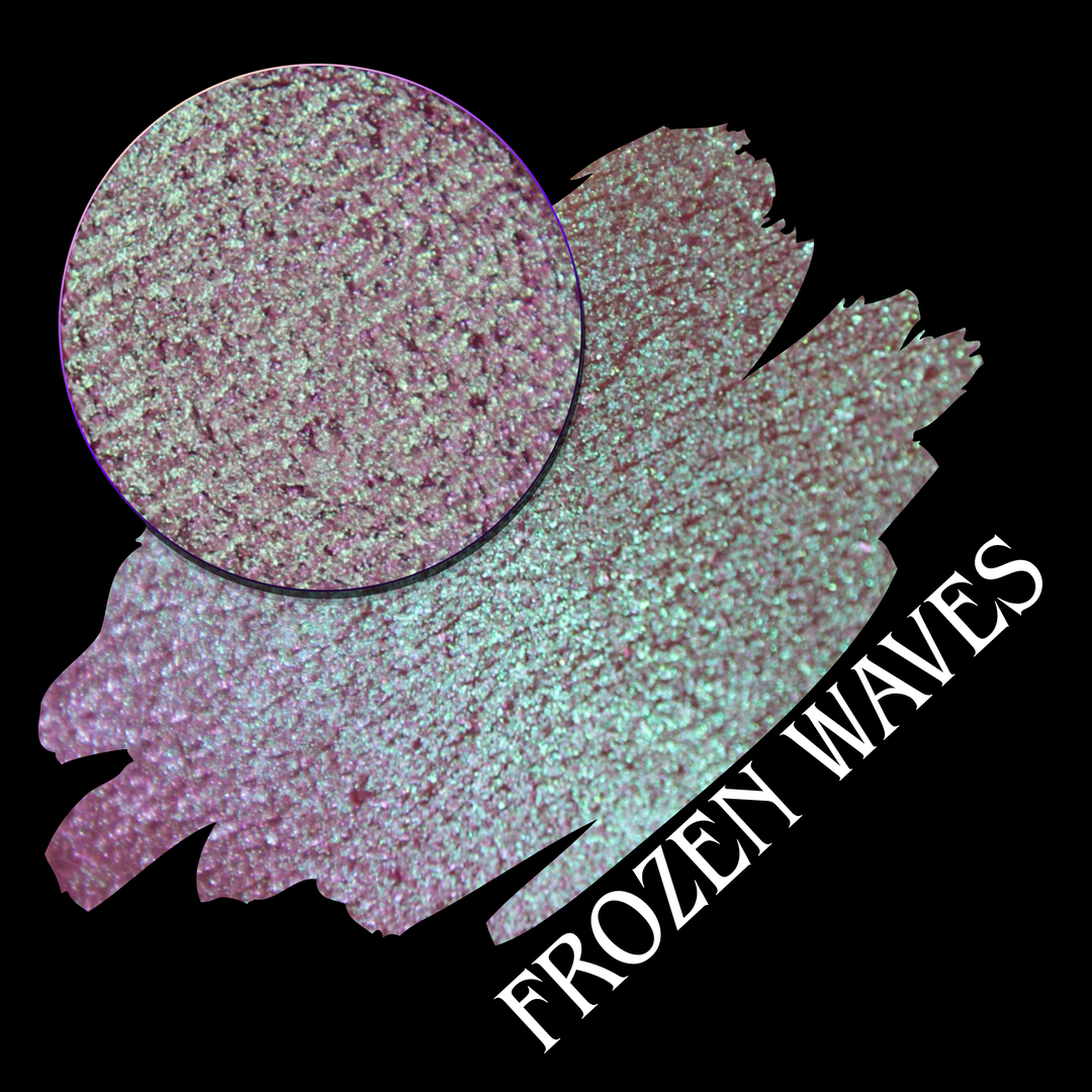 A round eyeshadow pan labeled 'FROZEN WAVES' made by Devinah Cosmetics, placed over a large crescent moon swatch of the same shimmering shifty eyeshadow on skin. The eyeshadow has a duochrome multichrome shift between colors red berry mauve teal green.