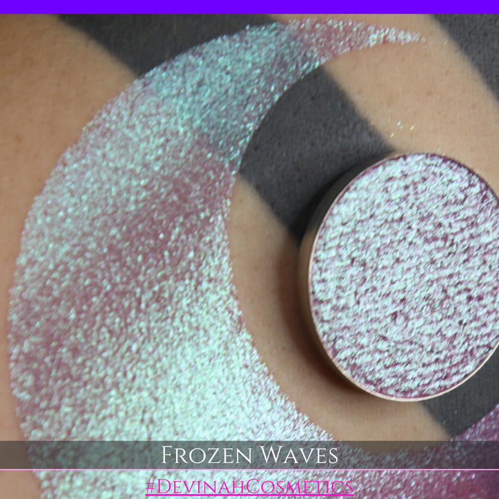 A round eyeshadow pan labeled 'FROZEN WAVES' made by Devinah Cosmetics, placed over a large crescent moon swatch of the same shimmering shifty eyeshadow on skin. The eyeshadow has a duochrome multichrome shift between colors red berry mauve teal green.