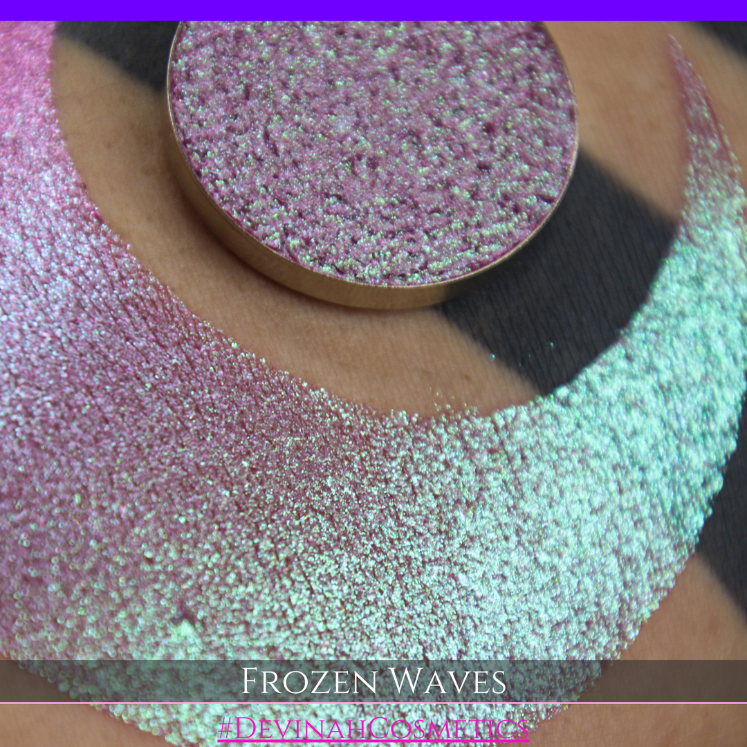 A round eyeshadow pan labeled 'FROZEN WAVES' made by Devinah Cosmetics, placed over a large crescent moon swatch of the same shimmering shifty eyeshadow on skin. The eyeshadow has a duochrome multichrome shift between colors red berry mauve teal green.