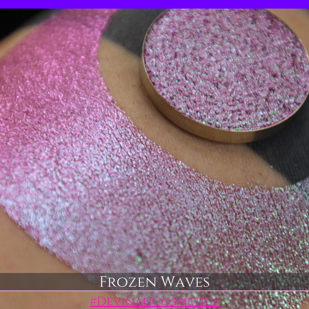 A round eyeshadow pan labeled 'FROZEN WAVES' made by Devinah Cosmetics, placed over a large crescent moon swatch of the same shimmering shifty eyeshadow on skin. The eyeshadow has a duochrome multichrome shift between colors red berry mauve teal green.