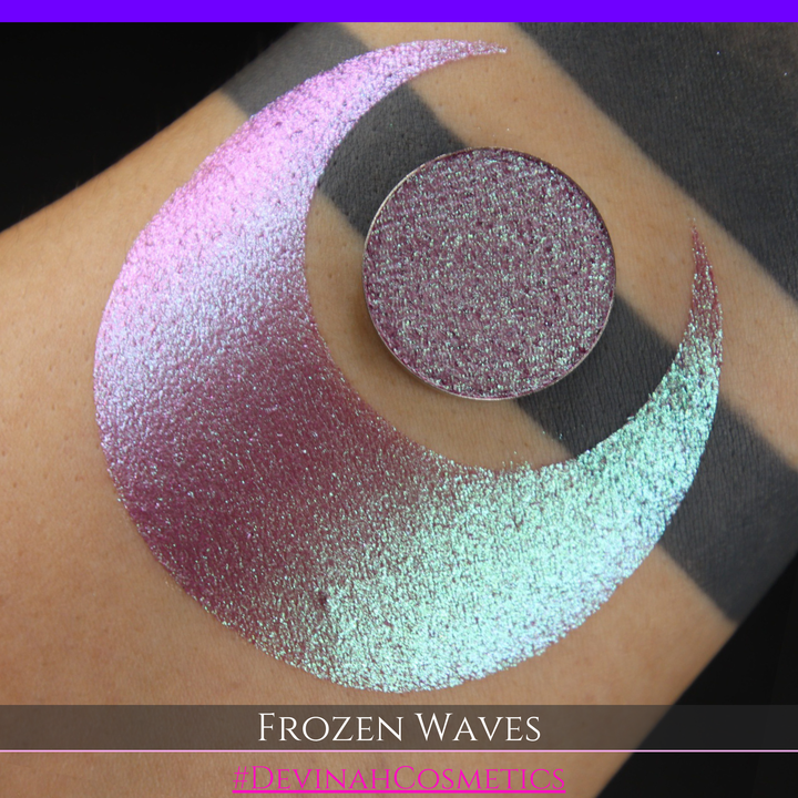 A round eyeshadow pan labeled 'FROZEN WAVES' made by Devinah Cosmetics, placed over a large crescent moon swatch of the same shimmering shifty eyeshadow on skin. The eyeshadow has a duochrome multichrome shift between colors red berry mauve teal green.
