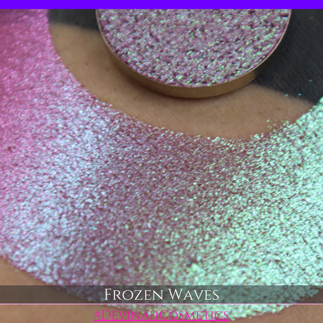 A round eyeshadow pan labeled 'FROZEN WAVES' made by Devinah Cosmetics, placed over a large crescent moon swatch of the same shimmering shifty eyeshadow on skin. The eyeshadow has a duochrome multichrome shift between colors red berry mauve teal green.