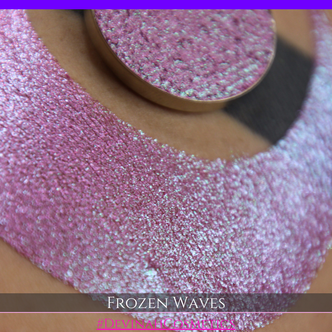 A round eyeshadow pan labeled 'FROZEN WAVES' made by Devinah Cosmetics, placed over a large crescent moon swatch of the same shimmering shifty eyeshadow on skin. The eyeshadow has a duochrome multichrome shift between colors red berry mauve teal green.