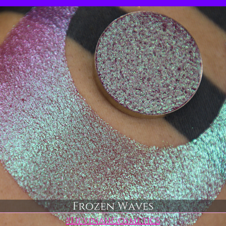 A round eyeshadow pan labeled 'FROZEN WAVES' made by Devinah Cosmetics, placed over a large crescent moon swatch of the same shimmering shifty eyeshadow on skin. The eyeshadow has a duochrome multichrome shift between colors red berry mauve teal green.