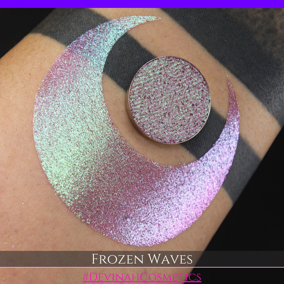 A round eyeshadow pan labeled 'FROZEN WAVES' made by Devinah Cosmetics, placed over a large crescent moon swatch of the same shimmering shifty eyeshadow on skin. The eyeshadow has a duochrome multichrome shift between colors red berry mauve teal green.