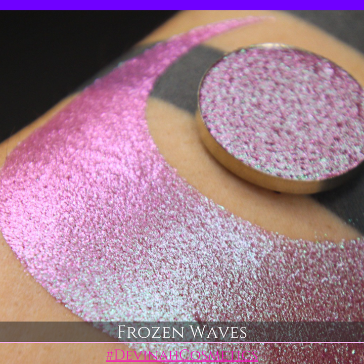 A round eyeshadow pan labeled 'FROZEN WAVES' made by Devinah Cosmetics, placed over a large crescent moon swatch of the same shimmering shifty eyeshadow on skin. The eyeshadow has a duochrome multichrome shift between colors red berry mauve teal green.