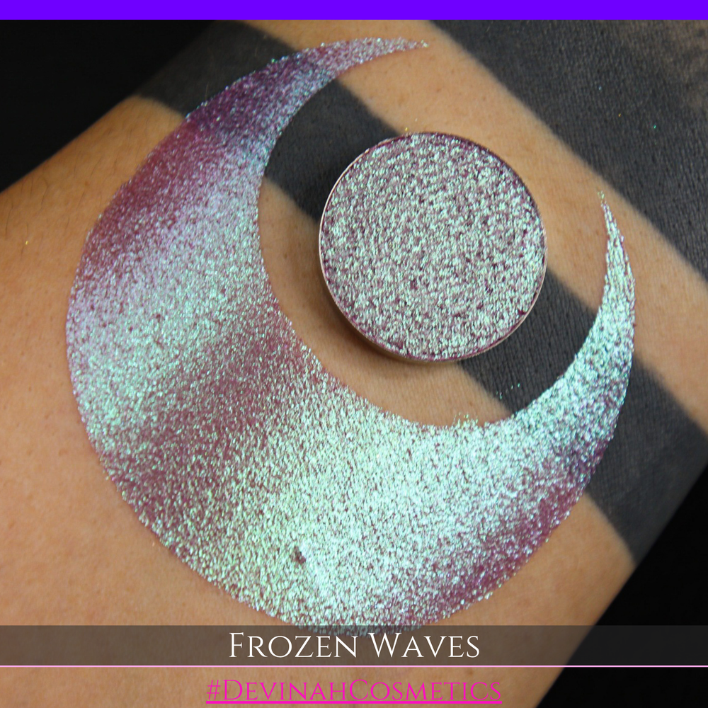 A round eyeshadow pan labeled 'FROZEN WAVES' made by Devinah Cosmetics, placed over a large crescent moon swatch of the same shimmering shifty eyeshadow on skin. The eyeshadow has a duochrome multichrome shift between colors red berry mauve teal green.