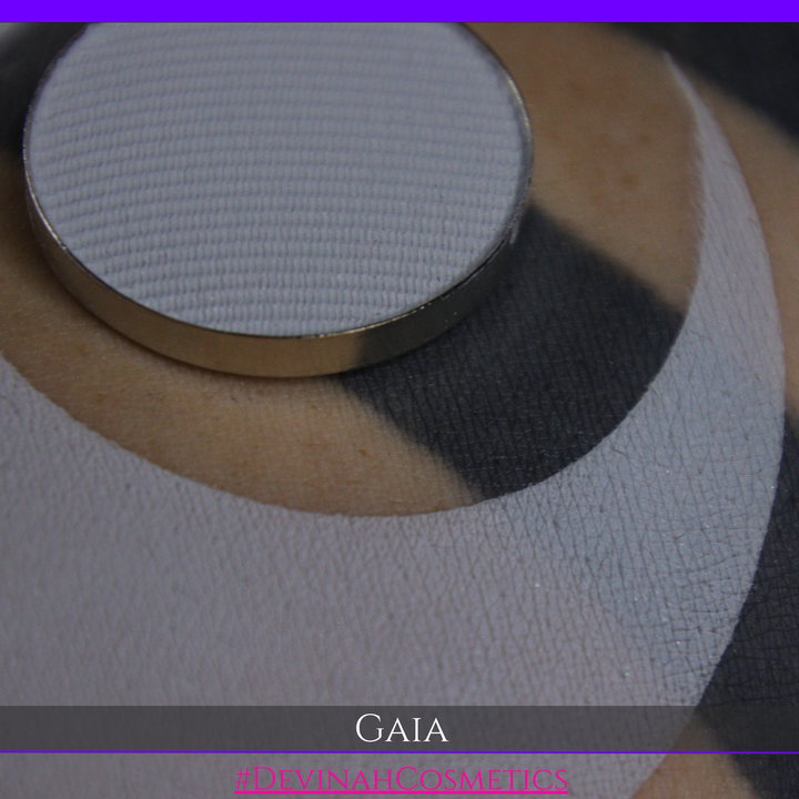 A close-up of a round makeup pan containing light gray powder, resting on a textured white and gray circular swatch. The brand name 'GAIA' and website 'DEVINAH COSMETICS' are displayed at the bottom.