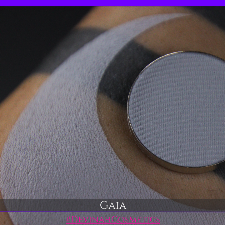 A close-up of a textured circular makeup palette pan labeled GAIA, resting on a beige-toned surface with a dark curved background.