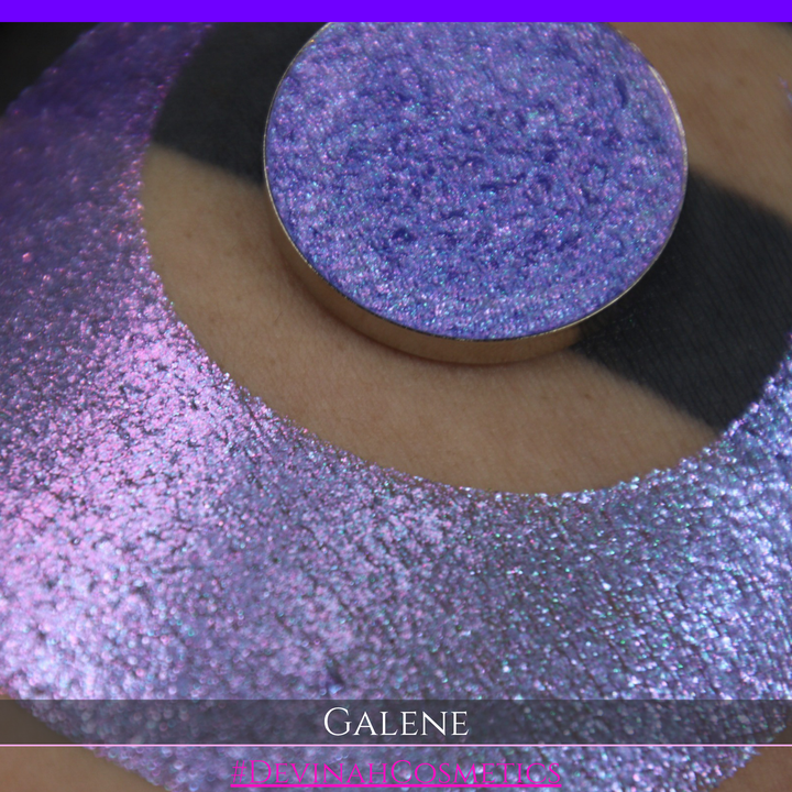 A round eyeshadow pan labeled 'GALENE' made by Devinah Cosmetics, placed over a large crescent moon swatch of the same shimmering shifty eyeshadow on skin. The duochorme multichrome eyeshadow has a shift between colors pink, purple, blue.