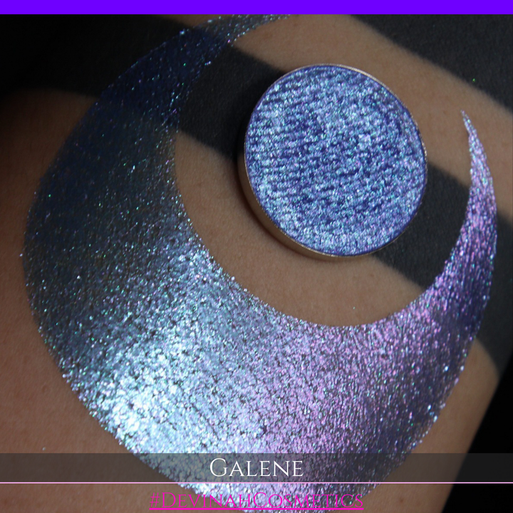 A round eyeshadow pan labeled 'GALENE' made by Devinah Cosmetics, placed over a large crescent moon swatch of the same shimmering shifty eyeshadow on skin. The duochorme multichrome eyeshadow has a shift between colors pink, purple, blue.