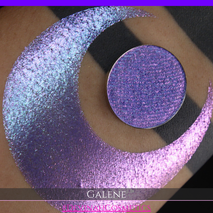 A round eyeshadow pan labeled 'GALENE' made by Devinah Cosmetics, placed over a large crescent moon swatch of the same shimmering shifty eyeshadow on skin. The duochorme multichrome eyeshadow has a shift between colors pink, purple, blue.