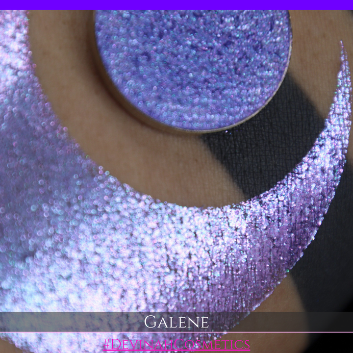 A round eyeshadow pan labeled 'GALENE' made by Devinah Cosmetics, placed over a large crescent moon swatch of the same shimmering shifty eyeshadow on skin. The duochorme multichrome eyeshadow has a shift between colors pink, purple, blue.