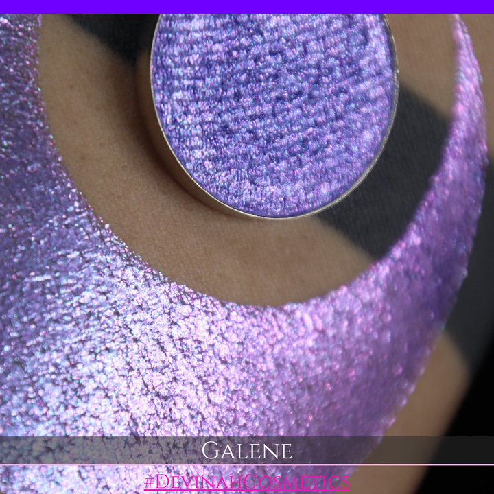 A round eyeshadow pan labeled 'GALENE' made by Devinah Cosmetics, placed over a large crescent moon swatch of the same shimmering shifty eyeshadow on skin. The duochorme multichrome eyeshadow has a shift between colors pink, purple, blue.