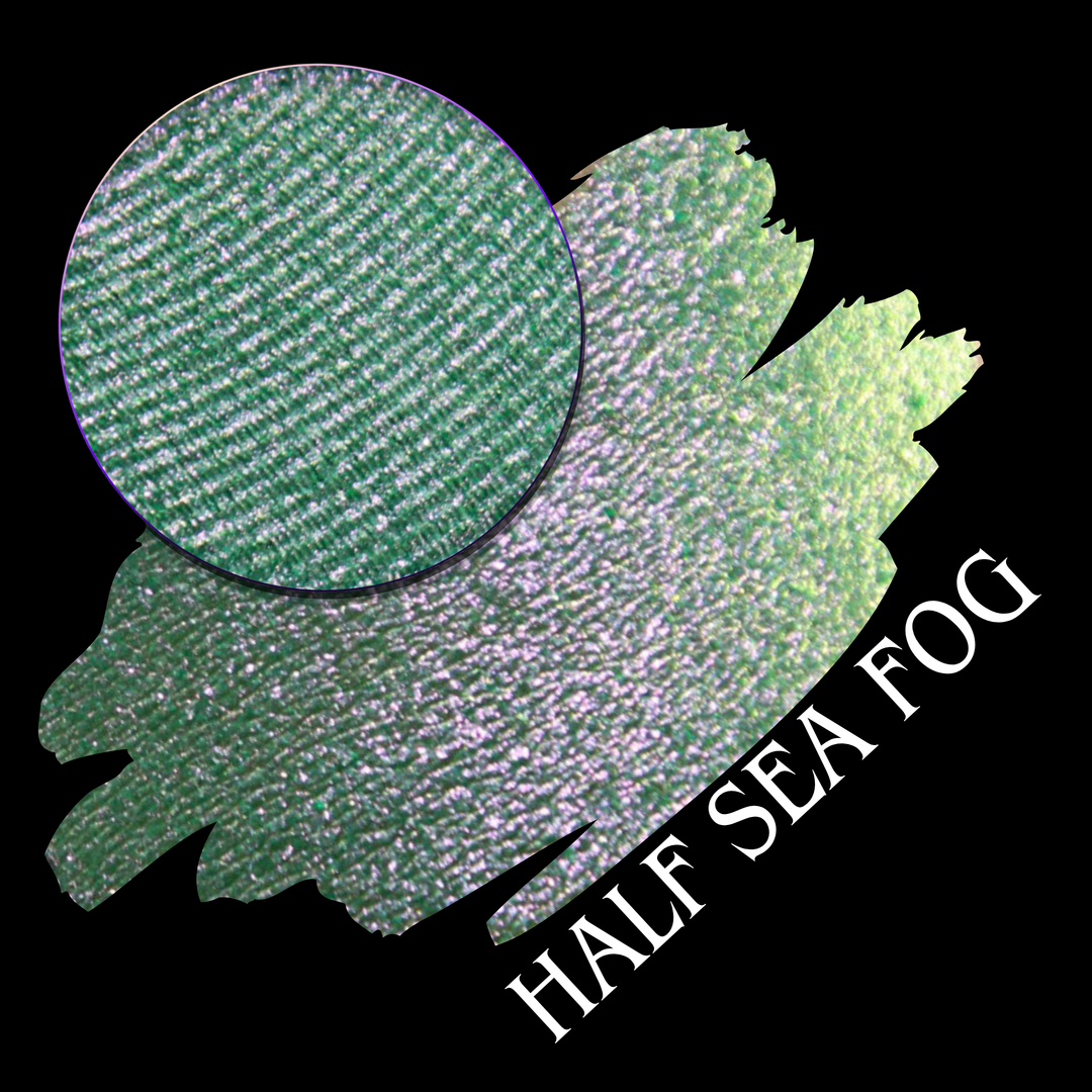 A round eyeshadow pan labeled 'HALF SEA FOG' made by Devinah Cosmetics, placed over a large crescent moon swatch of the same shimmering shifty eyeshadow on skin. The eyeshadow has a duochrome multichrome shift between colors lime green, yellow, pink.