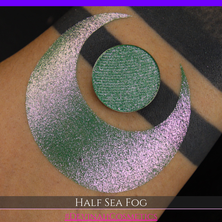 A round eyeshadow pan labeled 'HALF SEA FOG' made by Devinah Cosmetics, placed over a large crescent moon swatch of the same shimmering shifty eyeshadow on skin. The eyeshadow has a duochrome multichrome shift between colors lime green, yellow, pink.