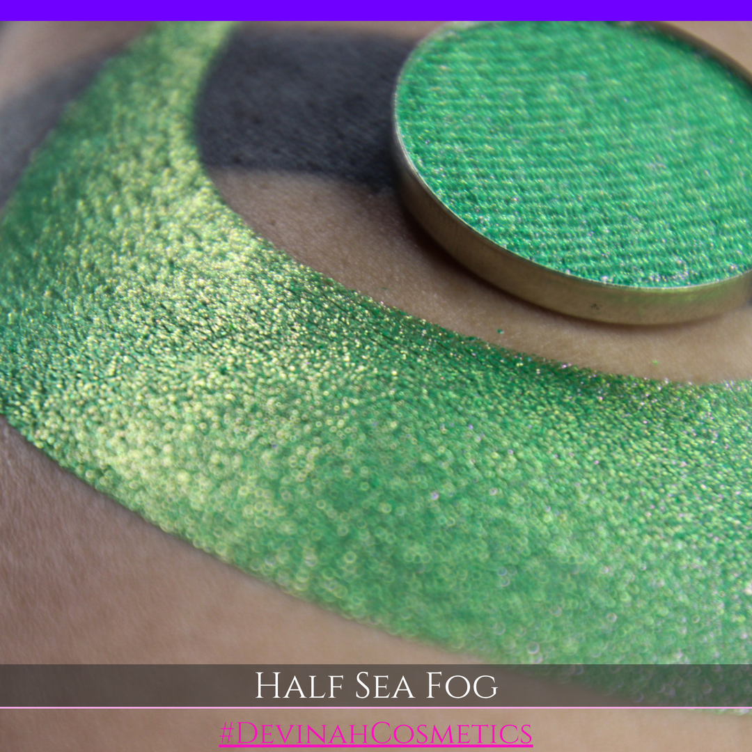 A round eyeshadow pan labeled 'HALF SEA FOG' made by Devinah Cosmetics, placed over a large crescent moon swatch of the same shimmering shifty eyeshadow on skin. The eyeshadow has a duochrome multichrome shift between colors lime green, yellow, pink.