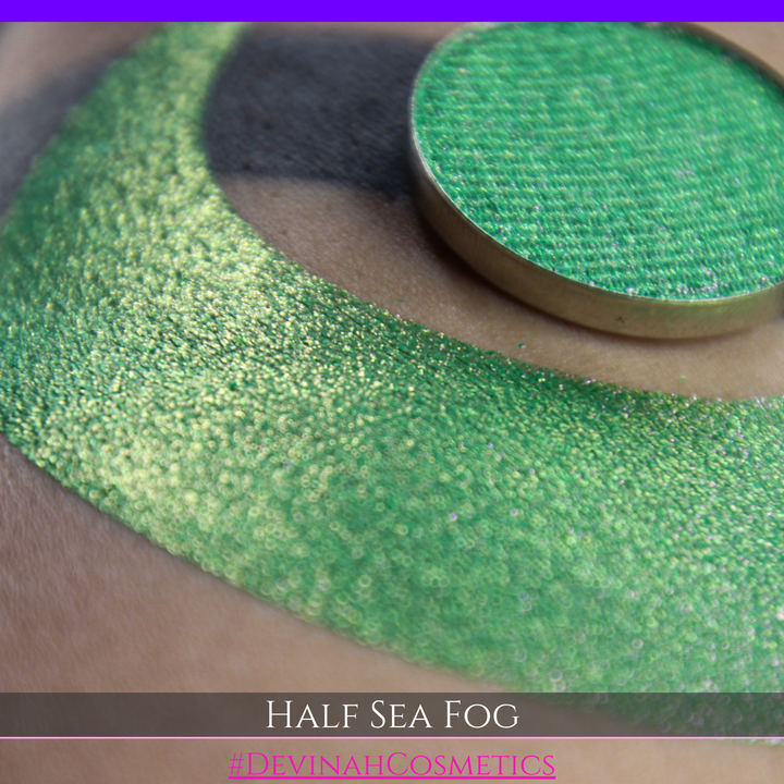 A round eyeshadow pan labeled 'HALF SEA FOG' made by Devinah Cosmetics, placed over a large crescent moon swatch of the same shimmering shifty eyeshadow on skin. The eyeshadow has a duochrome multichrome shift between colors lime green, yellow, pink.