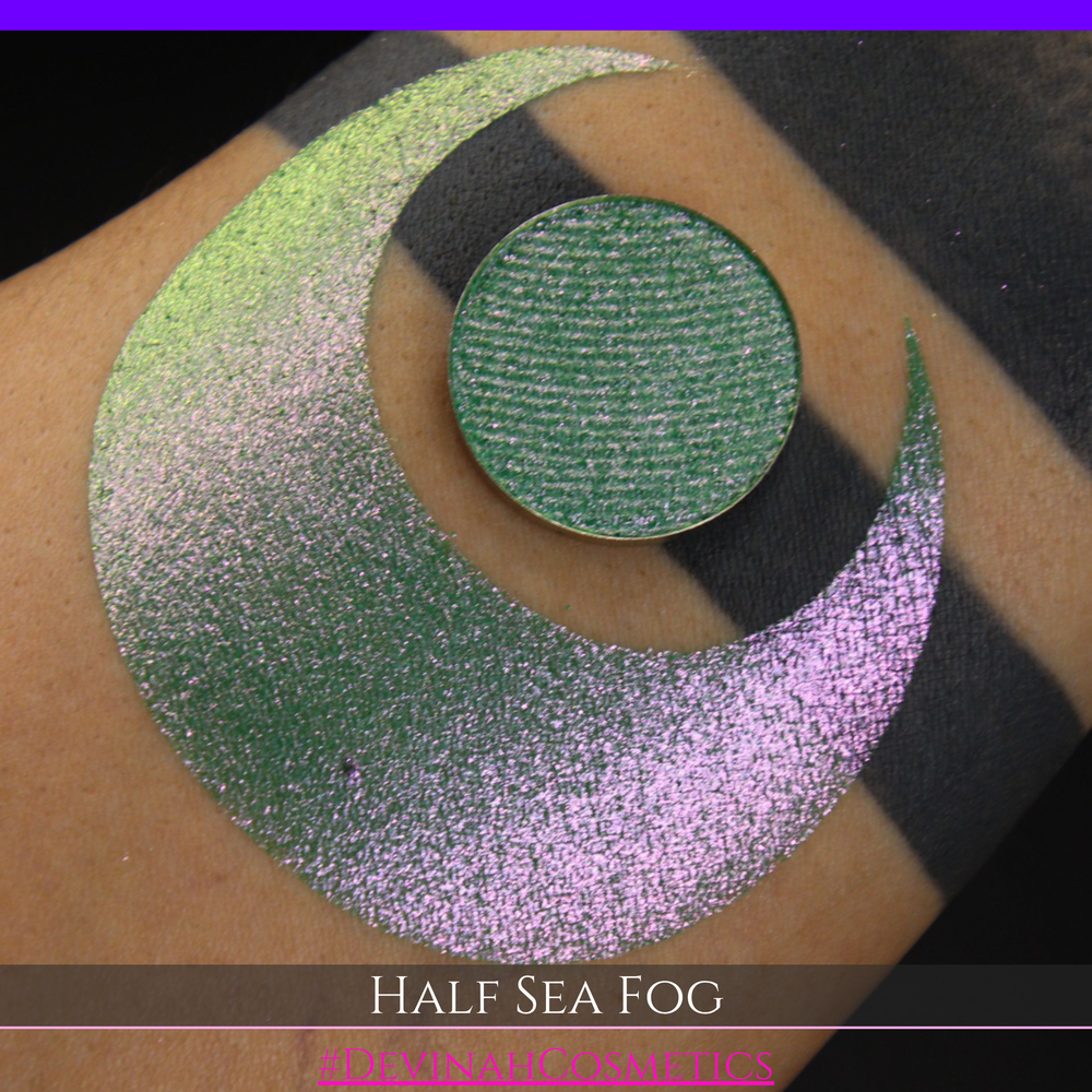 A round eyeshadow pan labeled 'HALF SEA FOG' made by Devinah Cosmetics, placed over a large crescent moon swatch of the same shimmering shifty eyeshadow on skin. The eyeshadow has a duochrome multichrome shift between colors lime green, yellow, pink.