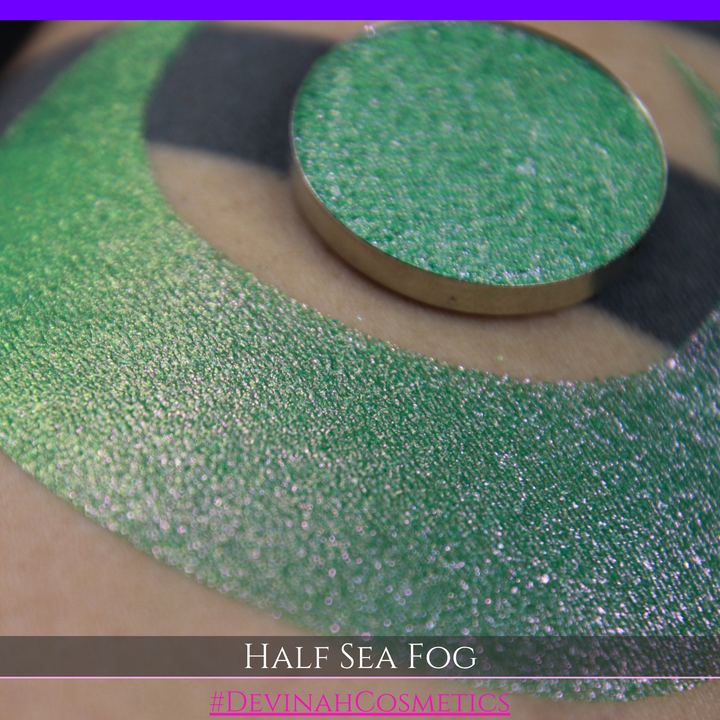 A round eyeshadow pan labeled 'HALF SEA FOG' made by Devinah Cosmetics, placed over a large crescent moon swatch of the same shimmering shifty eyeshadow on skin. The eyeshadow has a duochrome multichrome shift between colors lime green, yellow, pink.