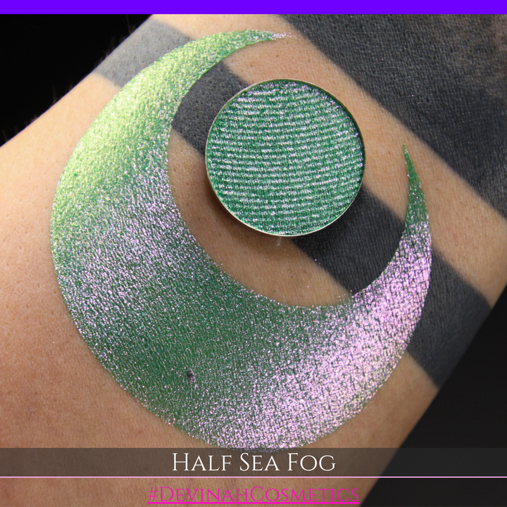 A round eyeshadow pan labeled 'HALF SEA FOG' made by Devinah Cosmetics, placed over a large crescent moon swatch of the same shimmering shifty eyeshadow on skin. The eyeshadow has a duochrome multichrome shift between colors lime green, yellow, pink.