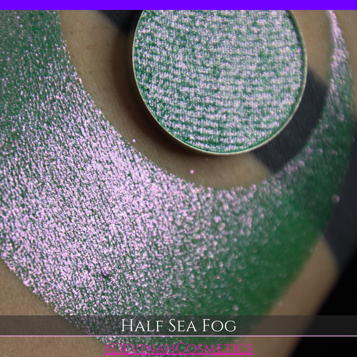 A round eyeshadow pan labeled 'HALF SEA FOG' made by Devinah Cosmetics, placed over a large crescent moon swatch of the same shimmering shifty eyeshadow on skin. The eyeshadow has a duochrome multichrome shift between colors lime green, yellow, pink.
