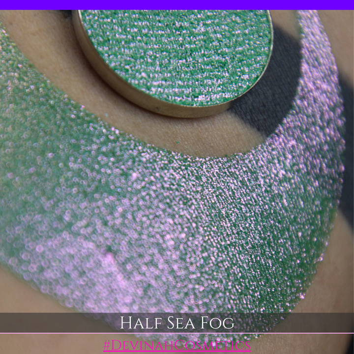 A round eyeshadow pan labeled 'HALF SEA FOG' made by Devinah Cosmetics, placed over a large crescent moon swatch of the same shimmering shifty eyeshadow on skin. The eyeshadow has a duochrome multichrome shift between colors lime green, yellow, pink.
